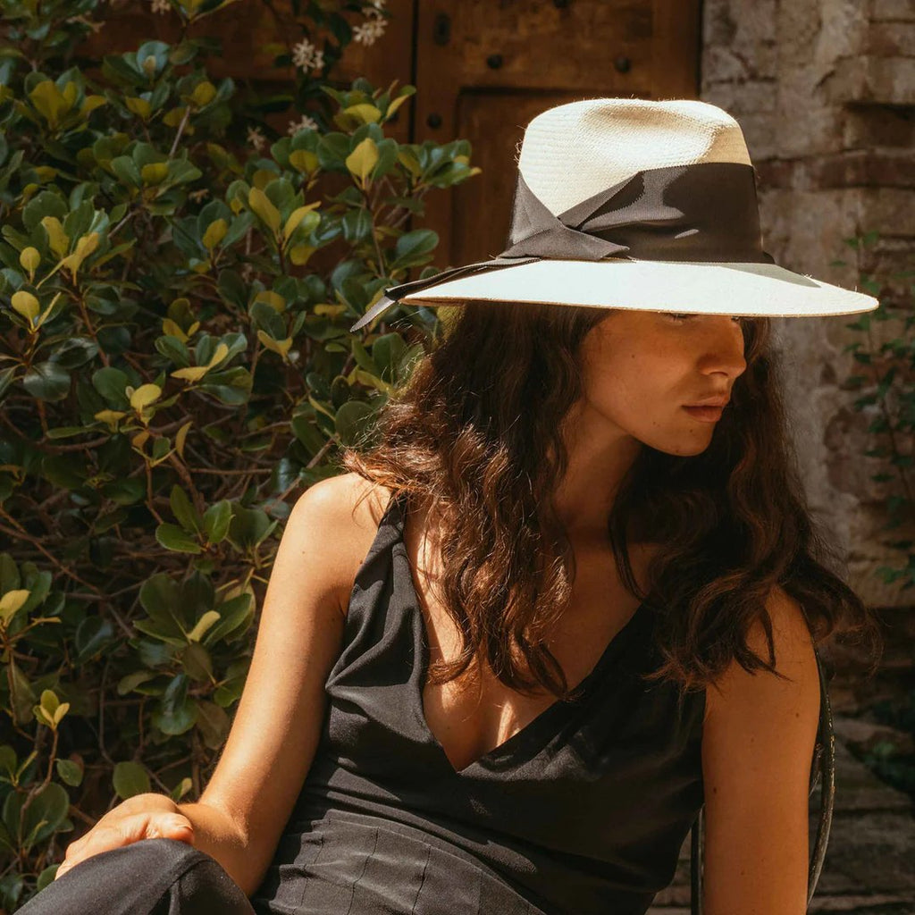 Women's Freya natural straw fedora with broad brim and oversized black grosgrain band, wrapped asymmetrically around the hat and finished with a modern angular fold, styled on model.