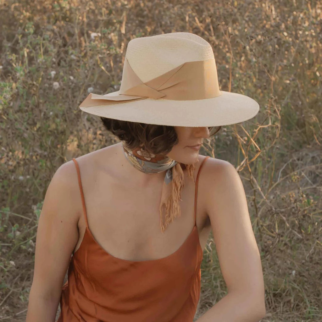 Freya women's tan straw fedora with broad brim and oversized honey grosgrain band, wrapped asymmetrically around the hat and finished with a modern angular fold styled on model. 
