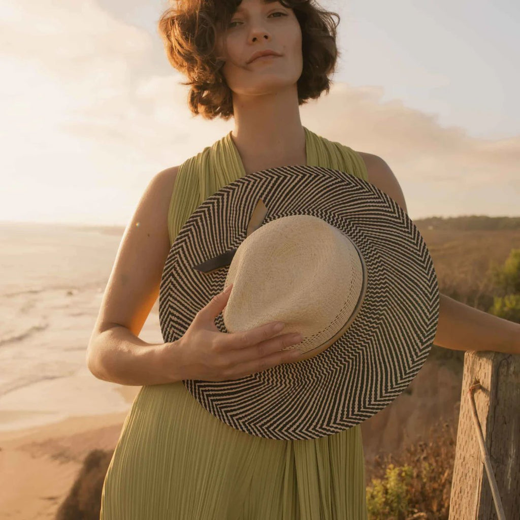 Freya Mesquite, vented natural crown paired with upturned cowgirl brim in black and natural, trimmed with hand-dyed cotton ribbon. Model is holding hat on a cliff by ocean at sunset.