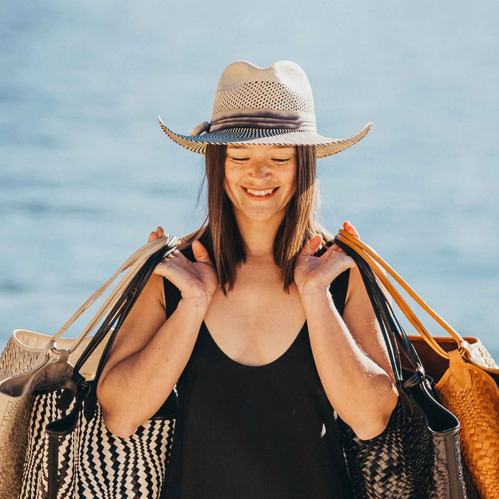 Freya Mesquite, vented natural crown paired with upturned cowgirl brim in black and natural, trimmed with hand-dyed cotton ribbon. Styled on smiling model who is holding bags in both arms. 